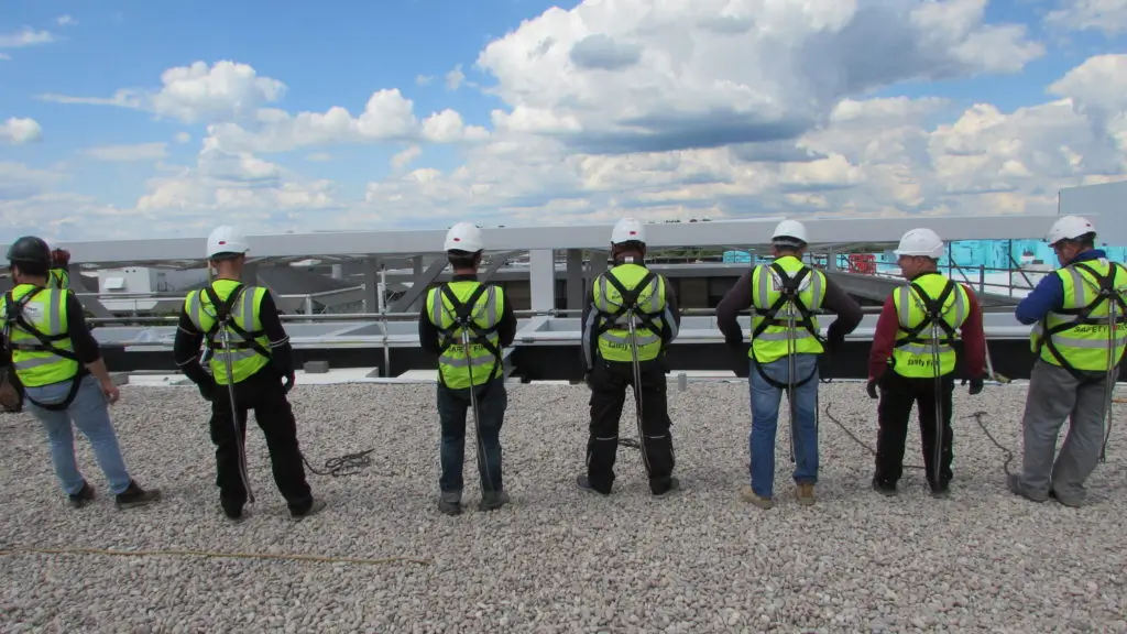 7 builders on height safety training course standing on the leading edge wearing yellow hi-vis vests, full body harness, lanyards and hard hats.