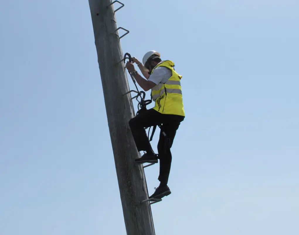 Worker climbing up pole wearing height safety equipment and yellow hi-vis vest.
